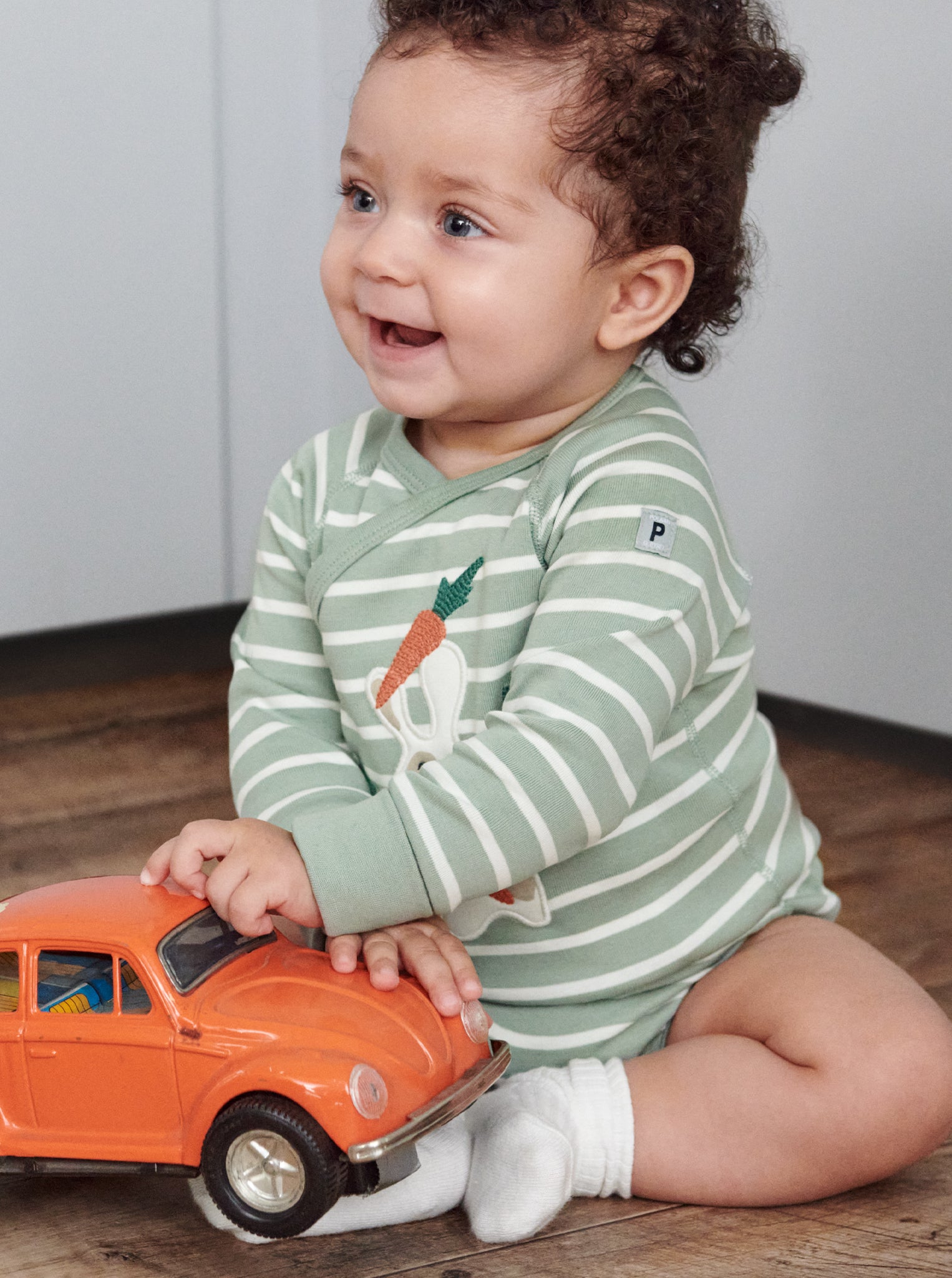 A toddler playing with orange car wearing a green and white striped cotton long sleeve babygrow with fun bunny eating carrots applique, paired with white organic cotton socks