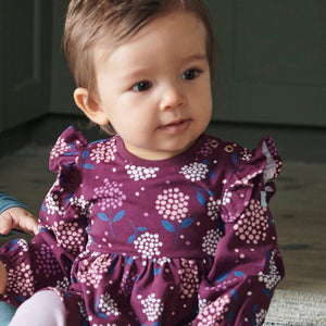 A close up of a baby girl wearing PO.P's sustainable purple floral baby dress made of organic cotton sitting on the floor