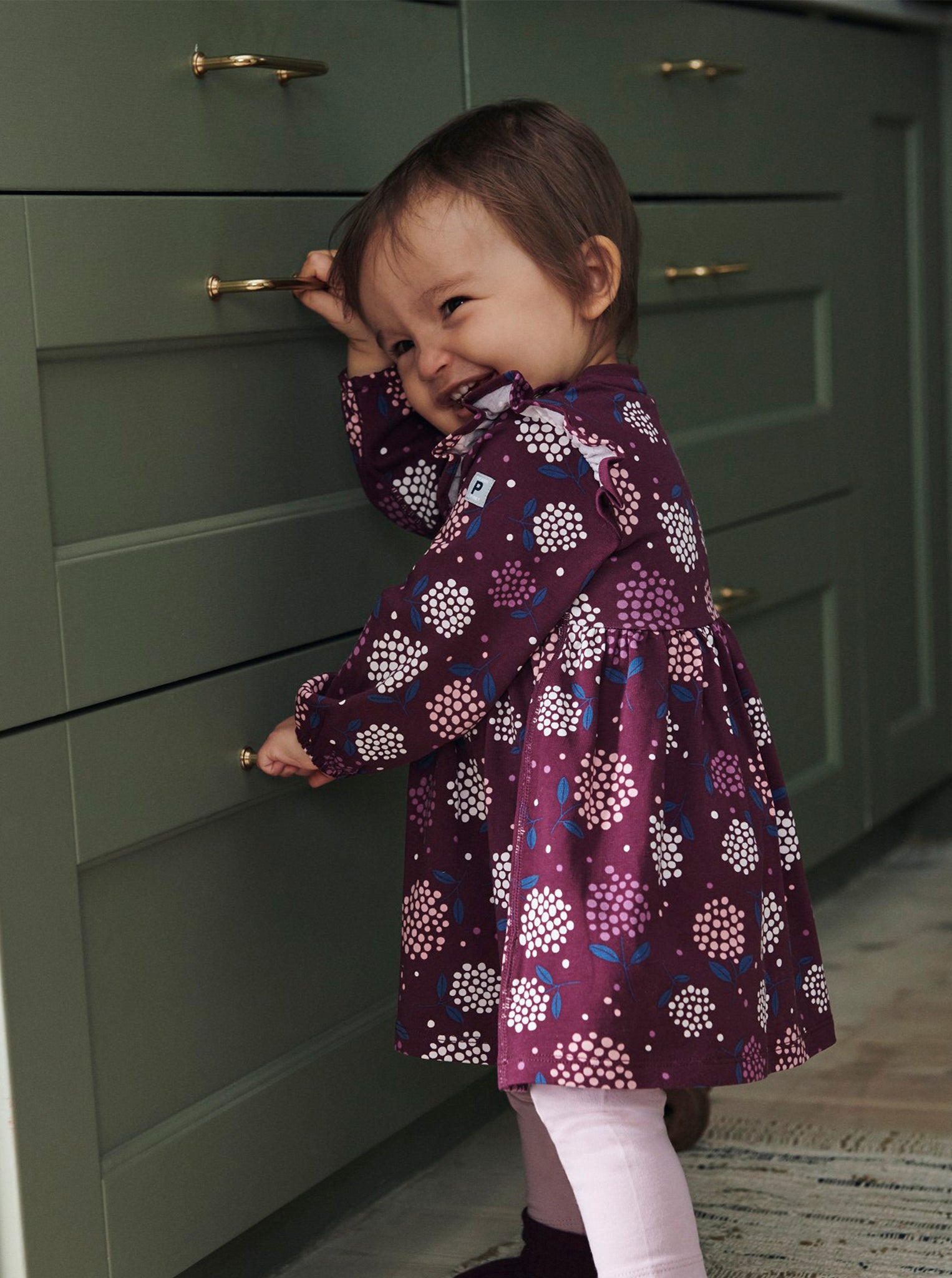 A baby girl wearing PO.P's sustainable purple floral baby dress made of organic cotton, playing with kitchen drawers