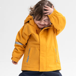 A happy little girl spotted wearing a yellow, kids waterproof jacket, with front pockets and reflectors, made of shell fabric.