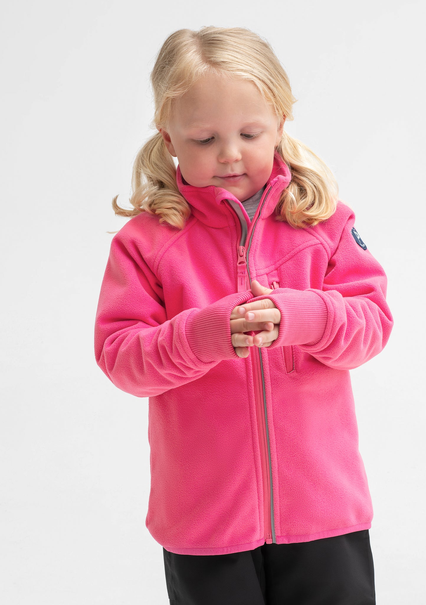 A young girl wearing a pink, kids waterproof fleece jacket, with reflector zips & cuff thumbholes, made of breathable fabric.