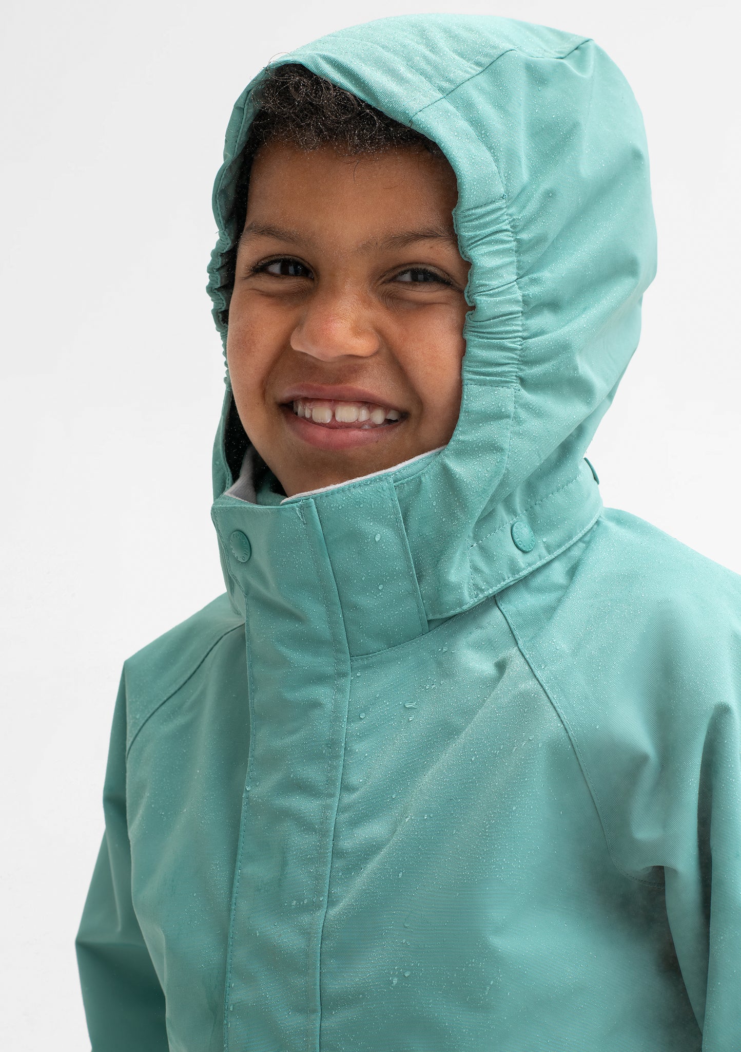 A boy smiling, wearing a green, hooded waterproof shell jacket, made of durable and lightweight fabric.